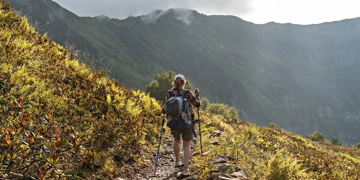 A woman with a backpack and trekking poles walks along a mountain trail surrounded by lush greenery and misty peaks in the distance.