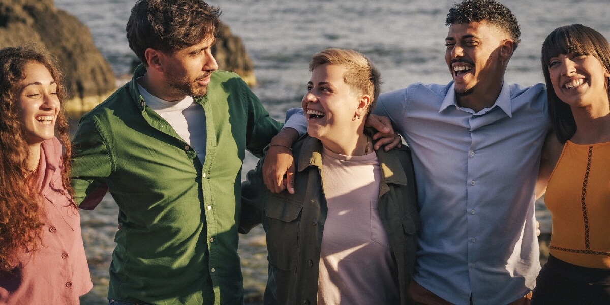 Group of five friends laughing together with arms around each other near a rocky shoreline.