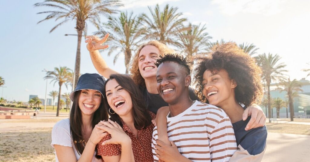 A group of friends laugh and embrace together outdoors on a sunny, palm-lined street.