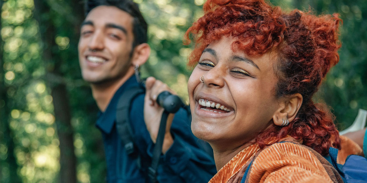 Two friends laugh together while hiking through a lush green forest with backpacks on.