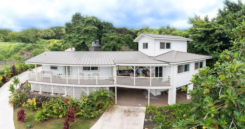 An aerial view of a large white house with a wraparound porch surrounded by lush tropical greenery.